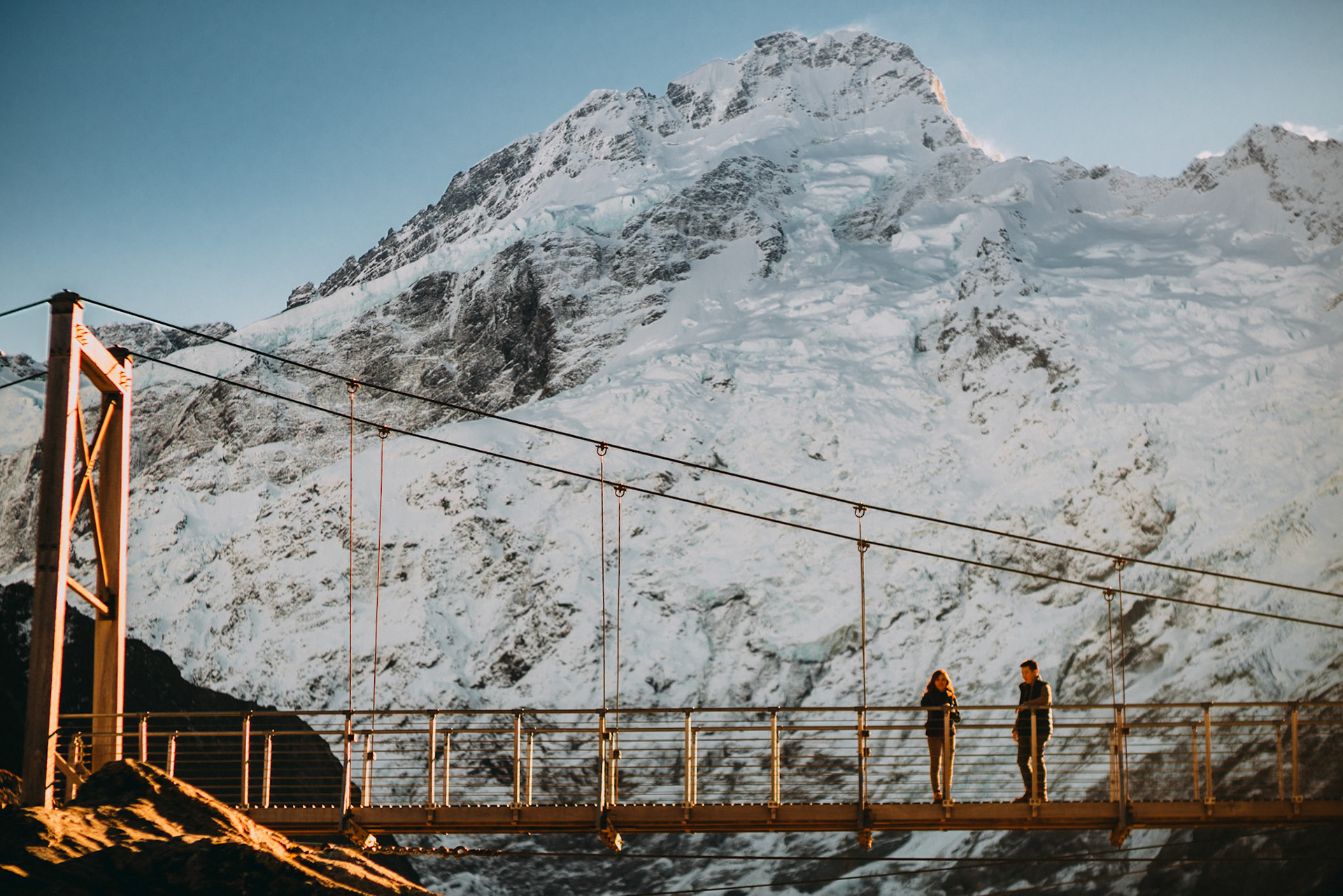 A couple on a bridge with a snowy mountain peak in the background, Aoraki Mount Cook National Park, New Zealand, June 2017, Sony A7RII.
