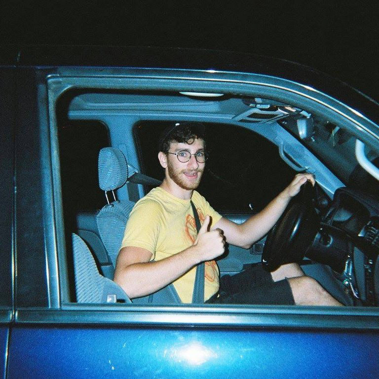 A grainy picture of a 20-something jewish man wearing jorts and a t-shirt with a boxing glove-wearing hotdog on it gives a thumbs up to the camera while sitting in the driver's seat of a 1995 Toyota Forerunner