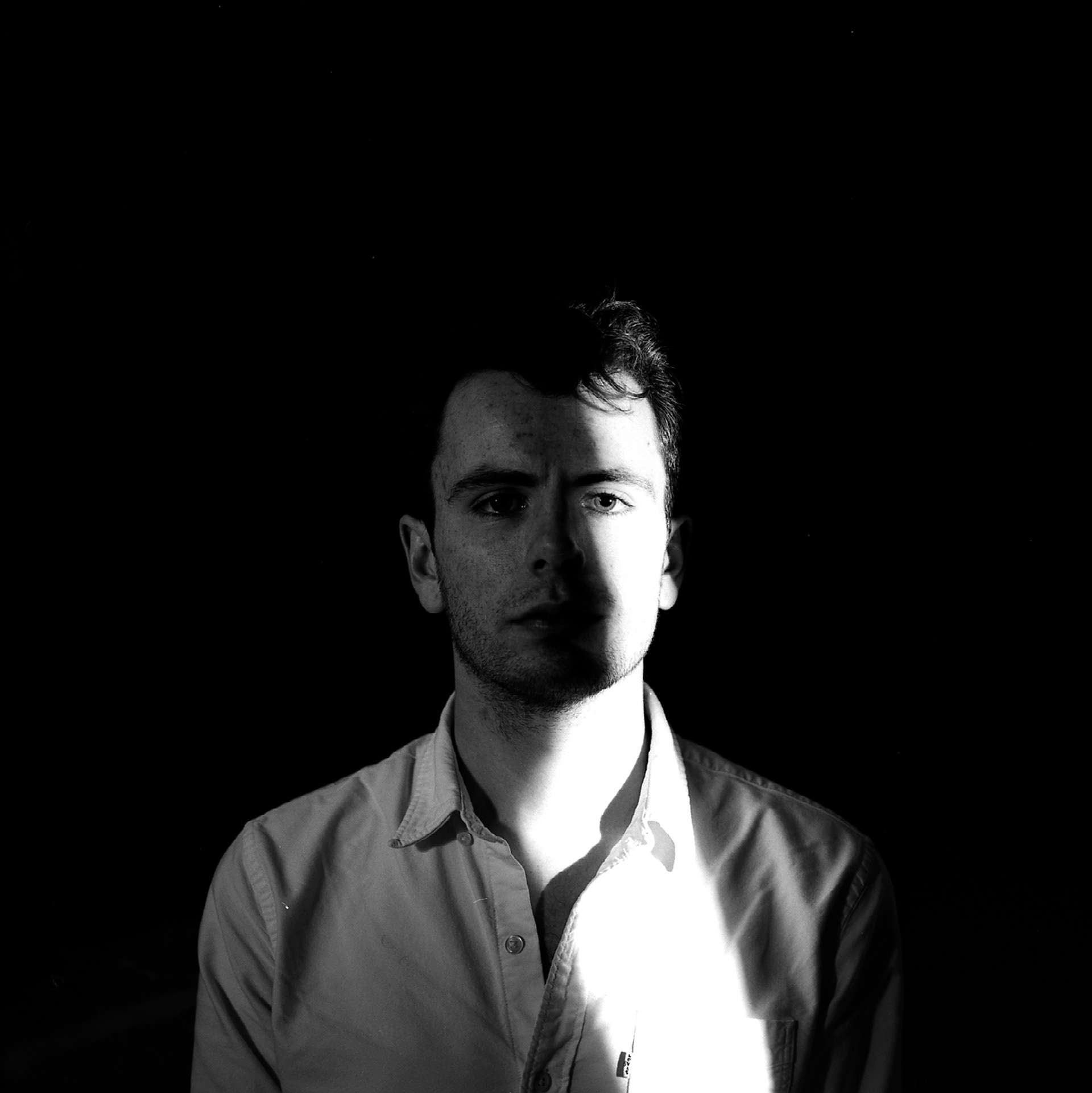 the male model looks directly into the camera, a small amount of his face is brightly lit, removing all detail, the rest is lit dimly