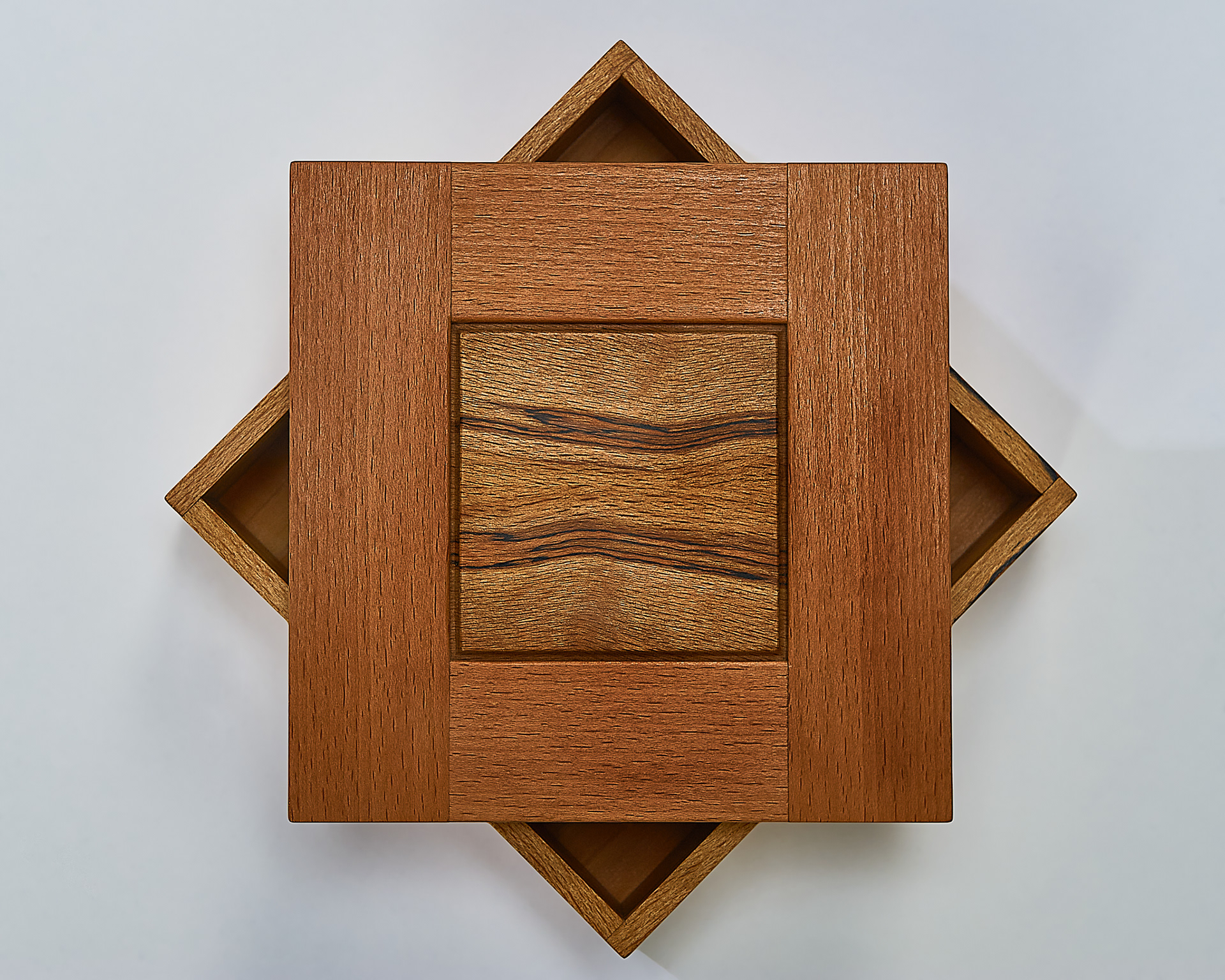 a top down view of a small, square wooden box. the lid is placed on top at an angle, showing some of the interior. the lid is a reddish wood, with patterned beech inlaid in the centre in a smaller square