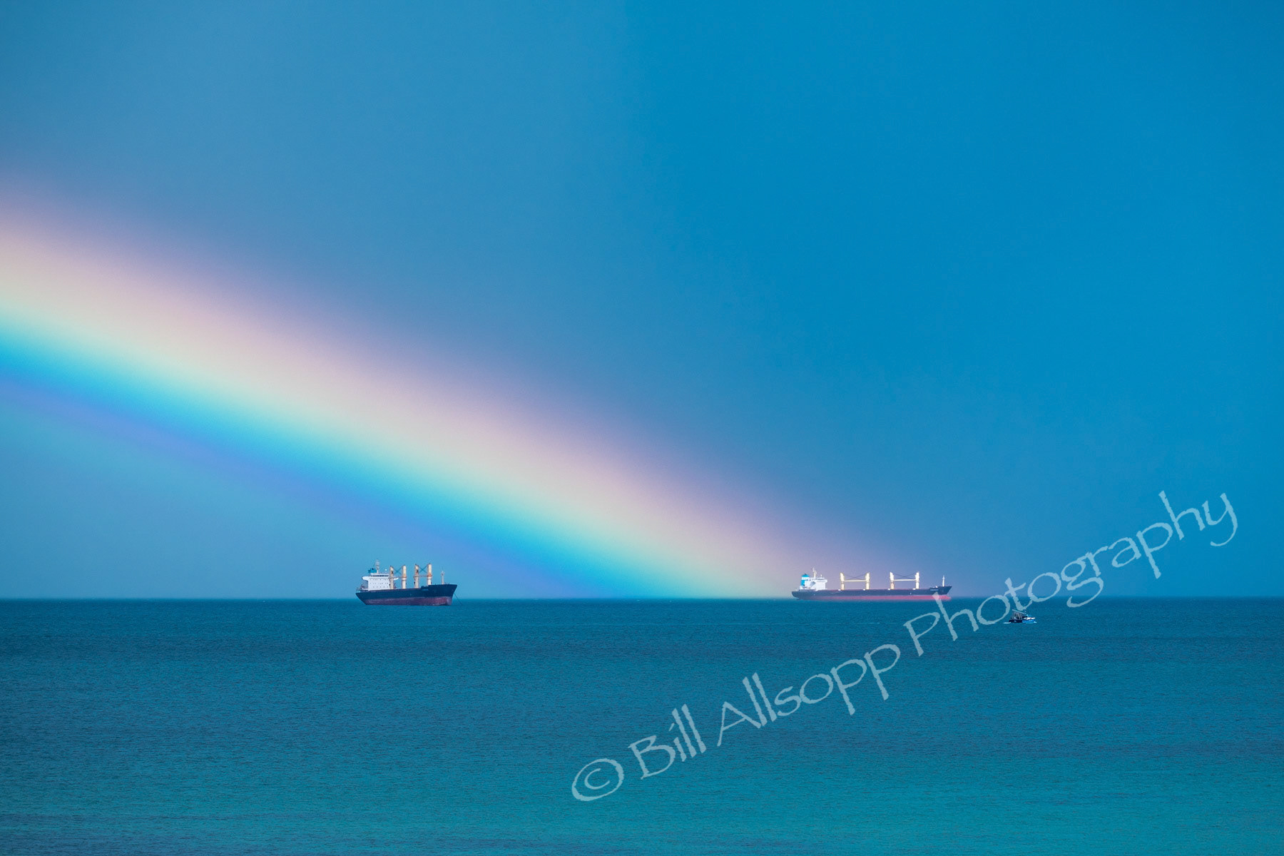 A rainbow at sea and two container ships at anchor.