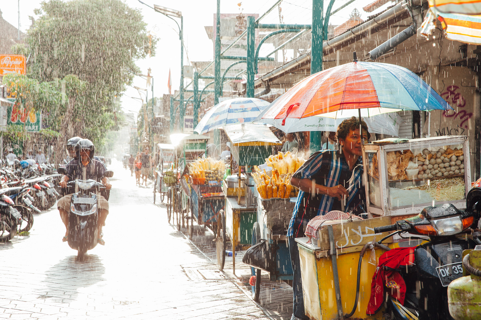 Indonesian food vendor hides from the rain under umbrella of his stall on the street of Kuta, Bali, Indonesia. Travel photography by Elena Ermakova.