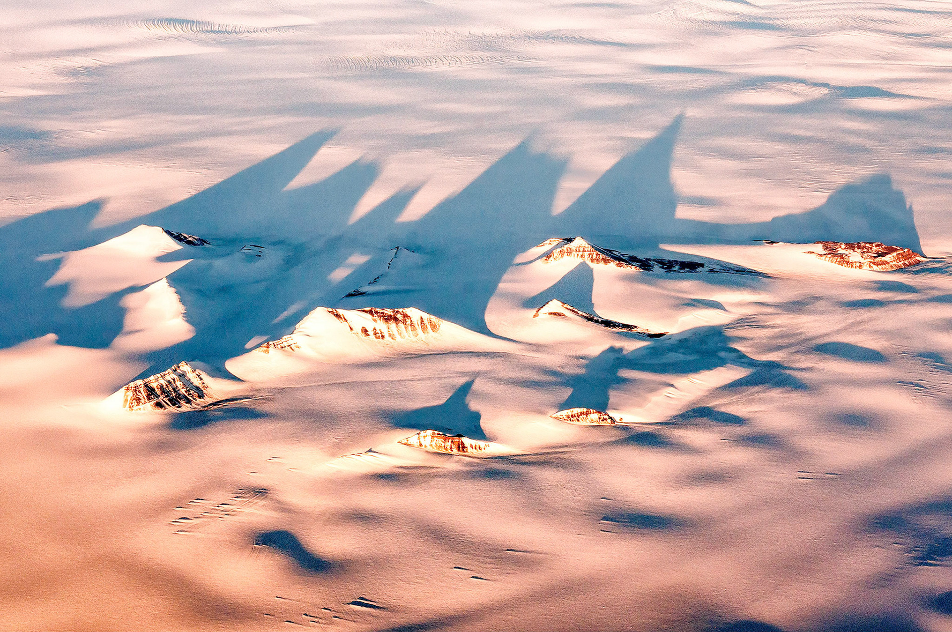 As the evening sun catches the tips of high mountain peaks in eastern Greenland, long shadows are cast over the vast, frigid landscape.