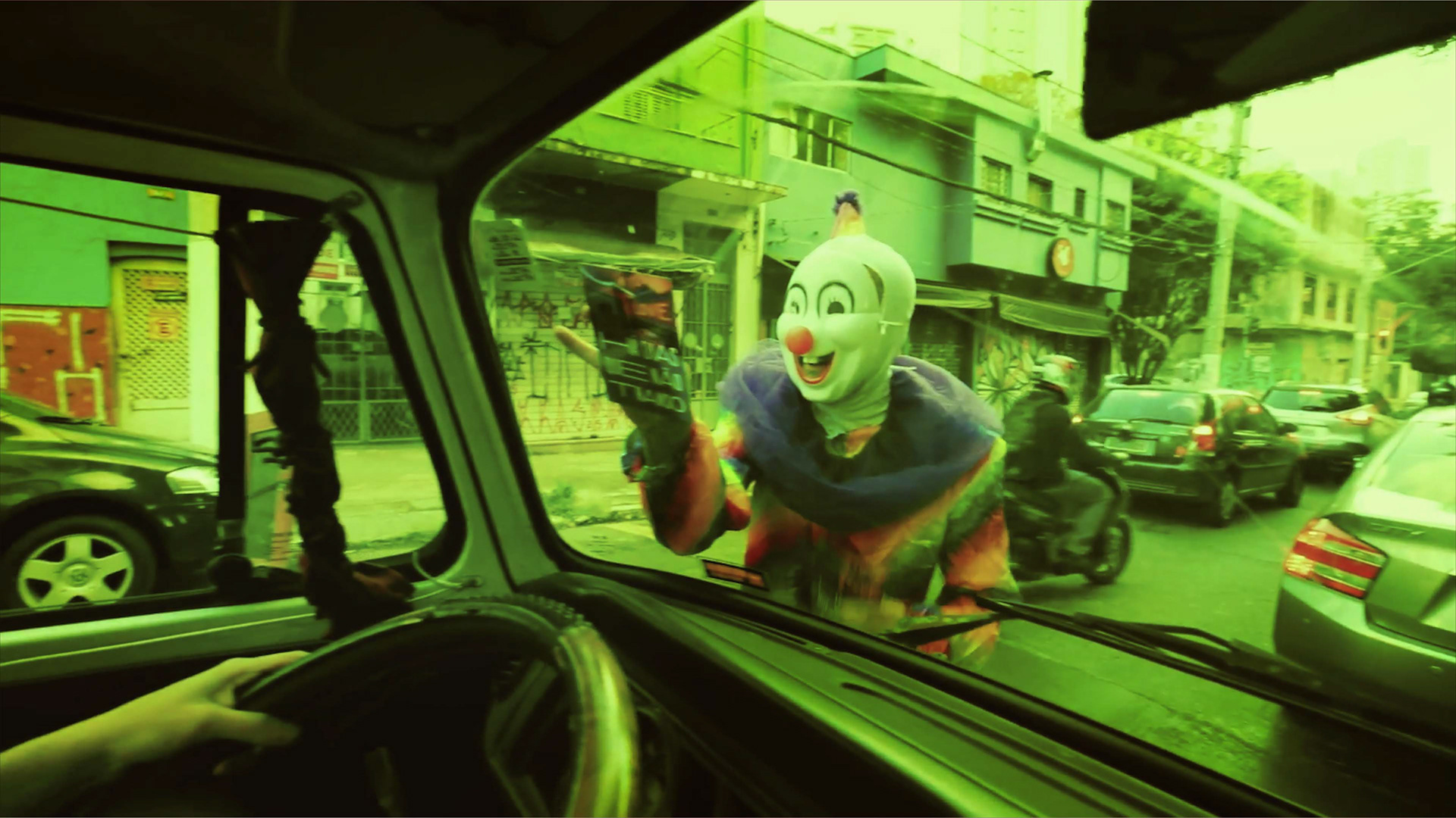 Uma foto com filtro esverdeada, mostra a visão de dentro de uma Kombi olhando para o vidro frontal. Bem no centro da imagem e pela rua, um palhaço de cara branca, nariz vermelho e roupa colorida coloca um flyer escrito "Há Dias Que não Transo". Ao fundo vemos muitos carros parados num semáforo fechado.
