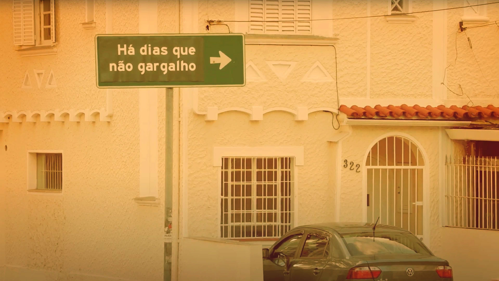 Uma casa amarela com detalhes brancos está na esquina de uma rua. Na frente e em destaque está uma placa de trânsito verde, bem grande, que em vez de sinalizar ruas ou indicações de trânsito, está apontando para a direita com o escrito "Há Dias Que Não Gargalho".