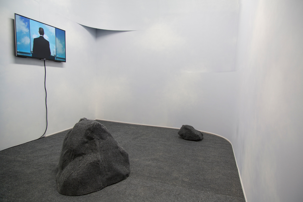 An installation shot of the project. Image description: a medium sizes room with a TV monitor hanging on the left wall and playing the short film "Man Above the Fog." At the corners of the room there are sections of the wall which are curved, with slits revealing the original corners of the room. The floor is covered with a grey carpet, and has boulder structures that are also made out of the same carpet. The boulders sat on motors which caused them to slowly rotate in the space. The room then, appears to mimic a kind of landscape.
