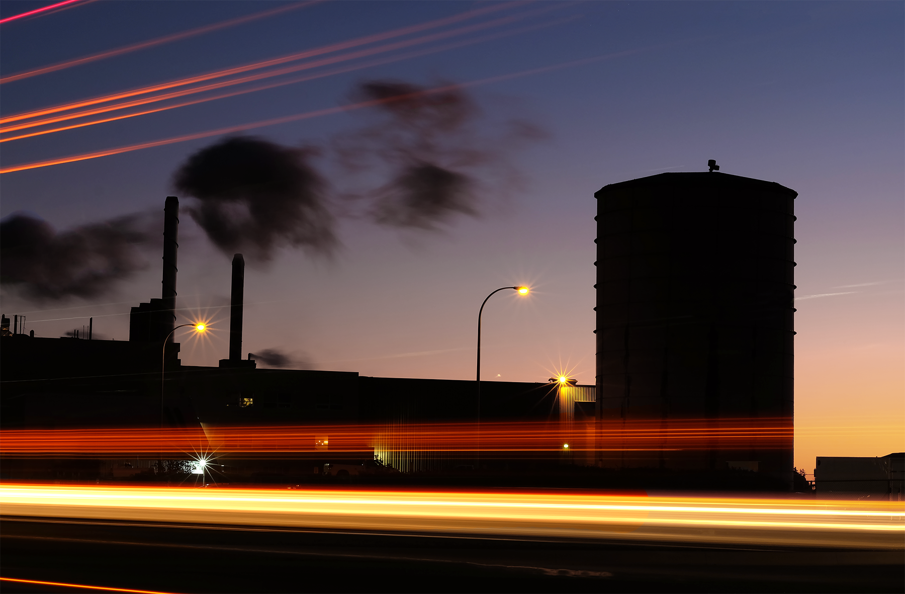 traffic streaks past a waste management plant at sunrise as dark smoke billows in the air